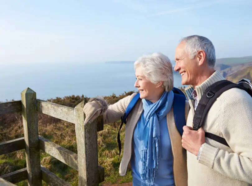 an older couple in gloves and scarves walking along a coastal path with the sea in the background